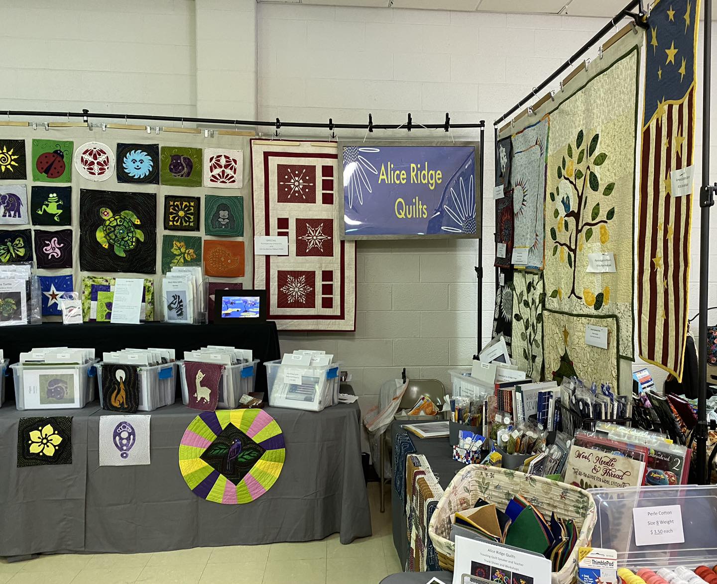 Booth display of Alice Ridge Quilts at the Heritage Quilters Guild show in Lenox, IL, showcasing various quilt patterns and kits, including designs of animals, flowers, and patriotic themes.